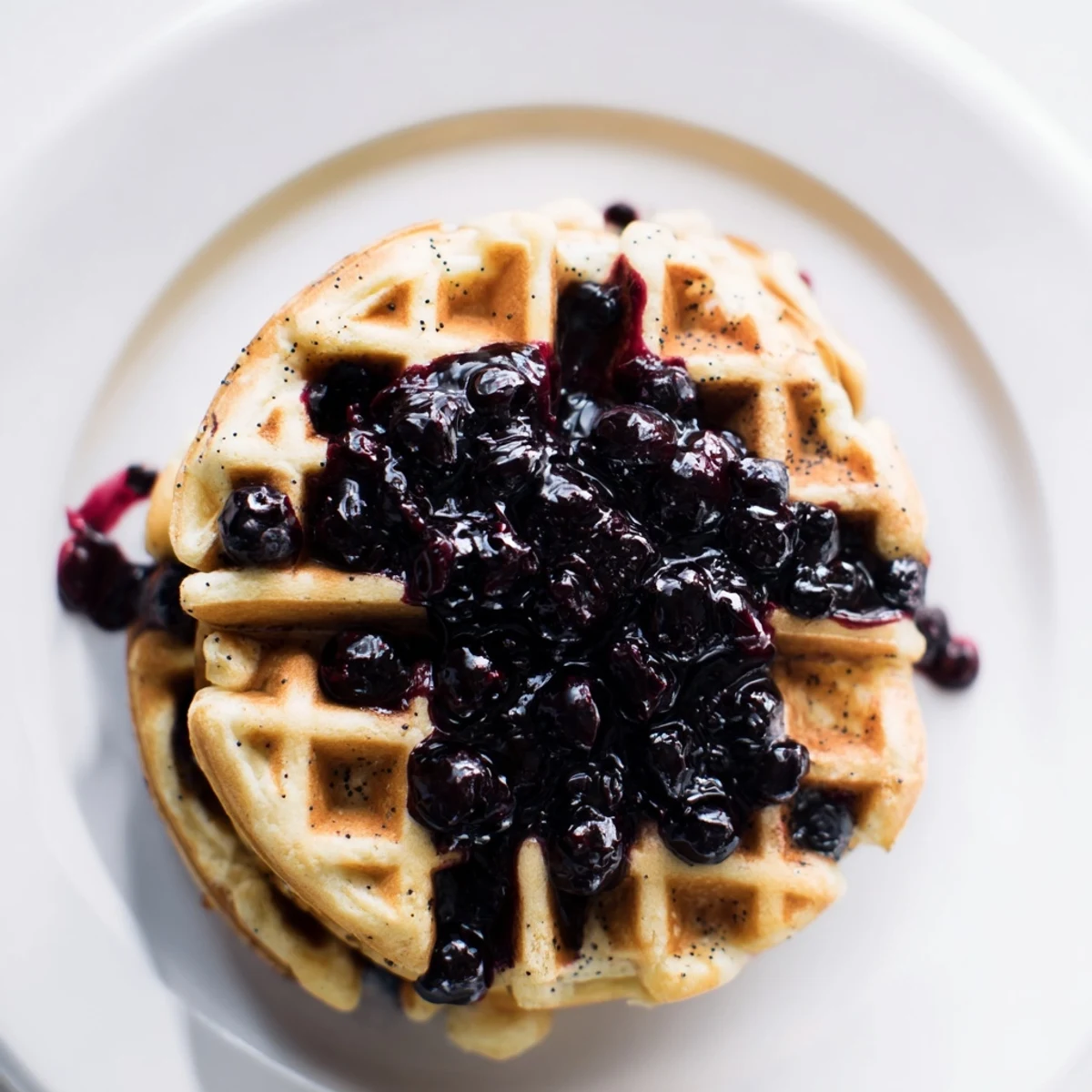 A close-up of golden crisp Lemon Poppy Seed Waffles drizzled with sweet blueberry compote and powdered sugar.