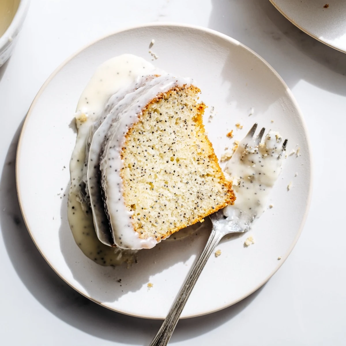 A golden-brown Lemon Poppy Seed Cake with a glossy lemon glaze is sliced on a white plate, showing a moist crumb with poppy seeds throughout.